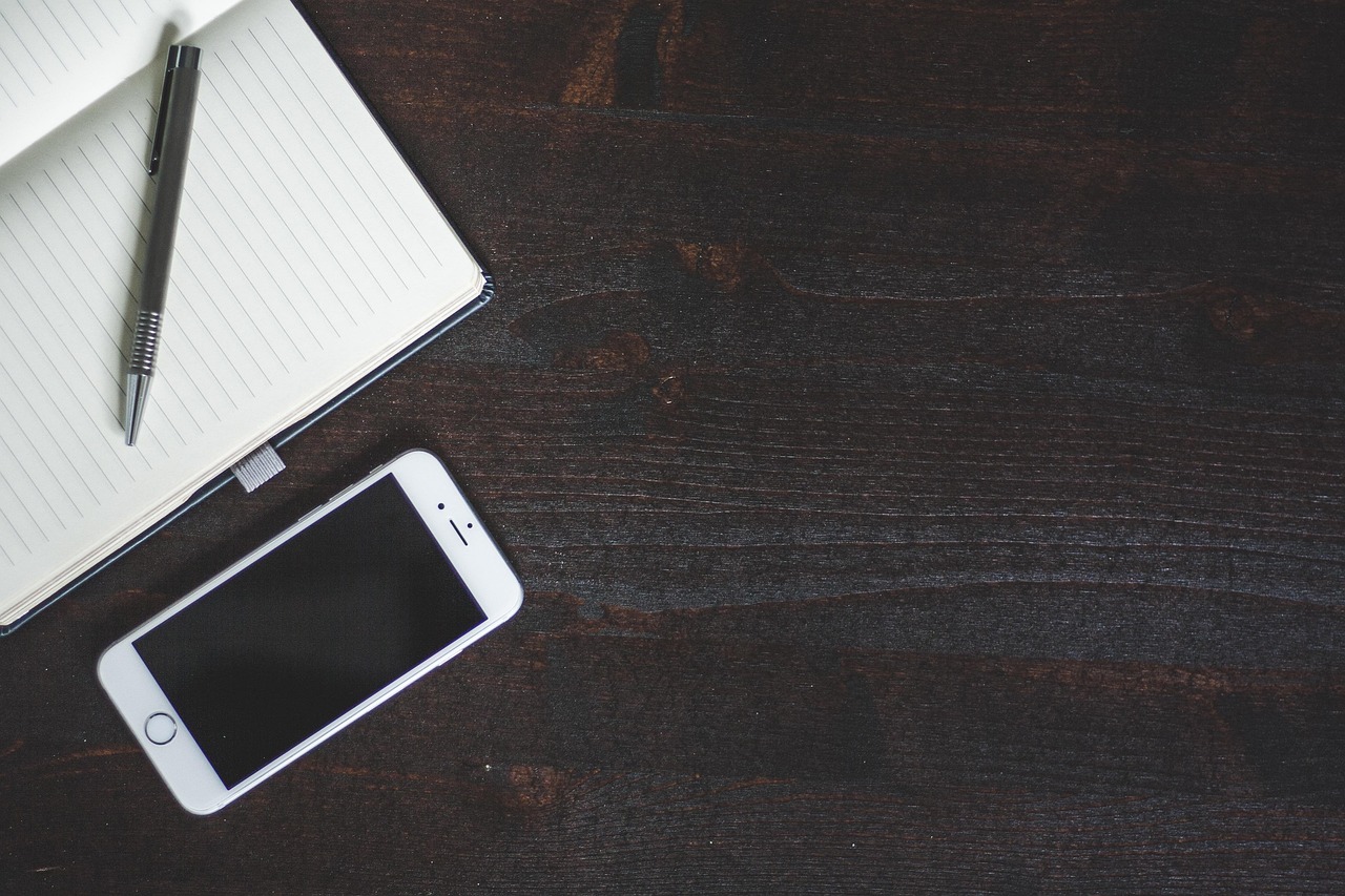 An iPhone, notebook, and pen laid out on a dark wood desk